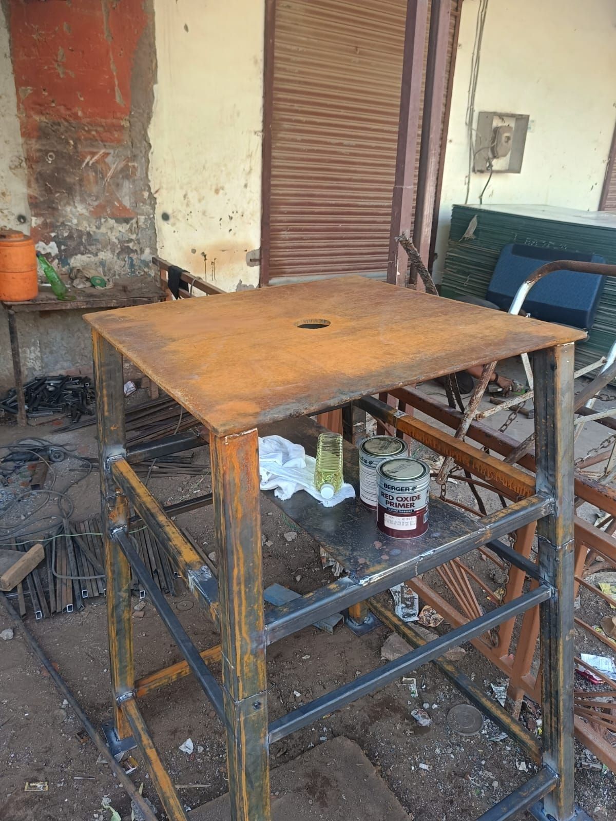 The welded steel stand outside the fabrication shop, tins of red-oxide primer on its lower shelf.