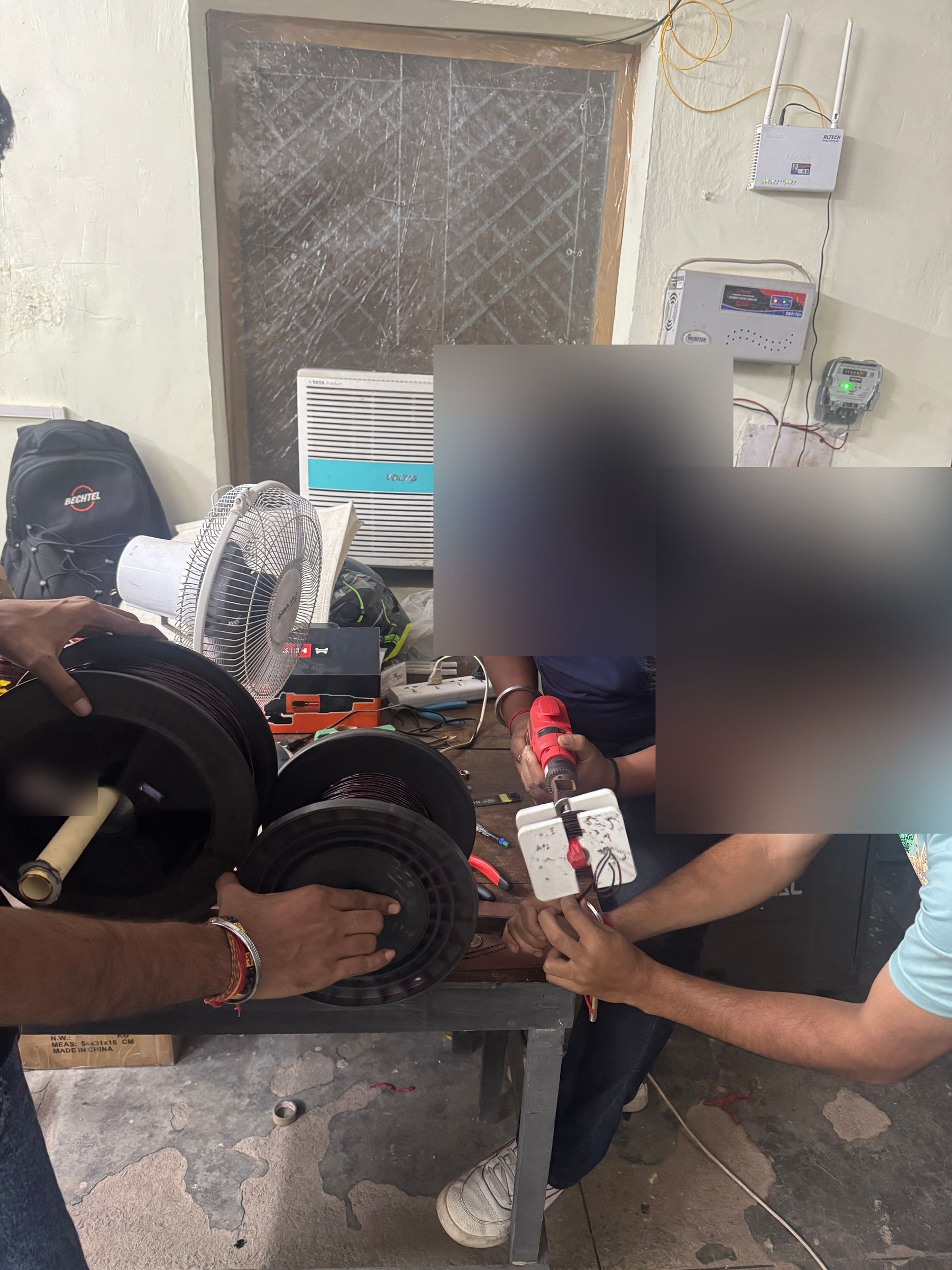 Two people at the bench winding a coil former with a hand drill, spools of copper wire held ready, faces blurred.