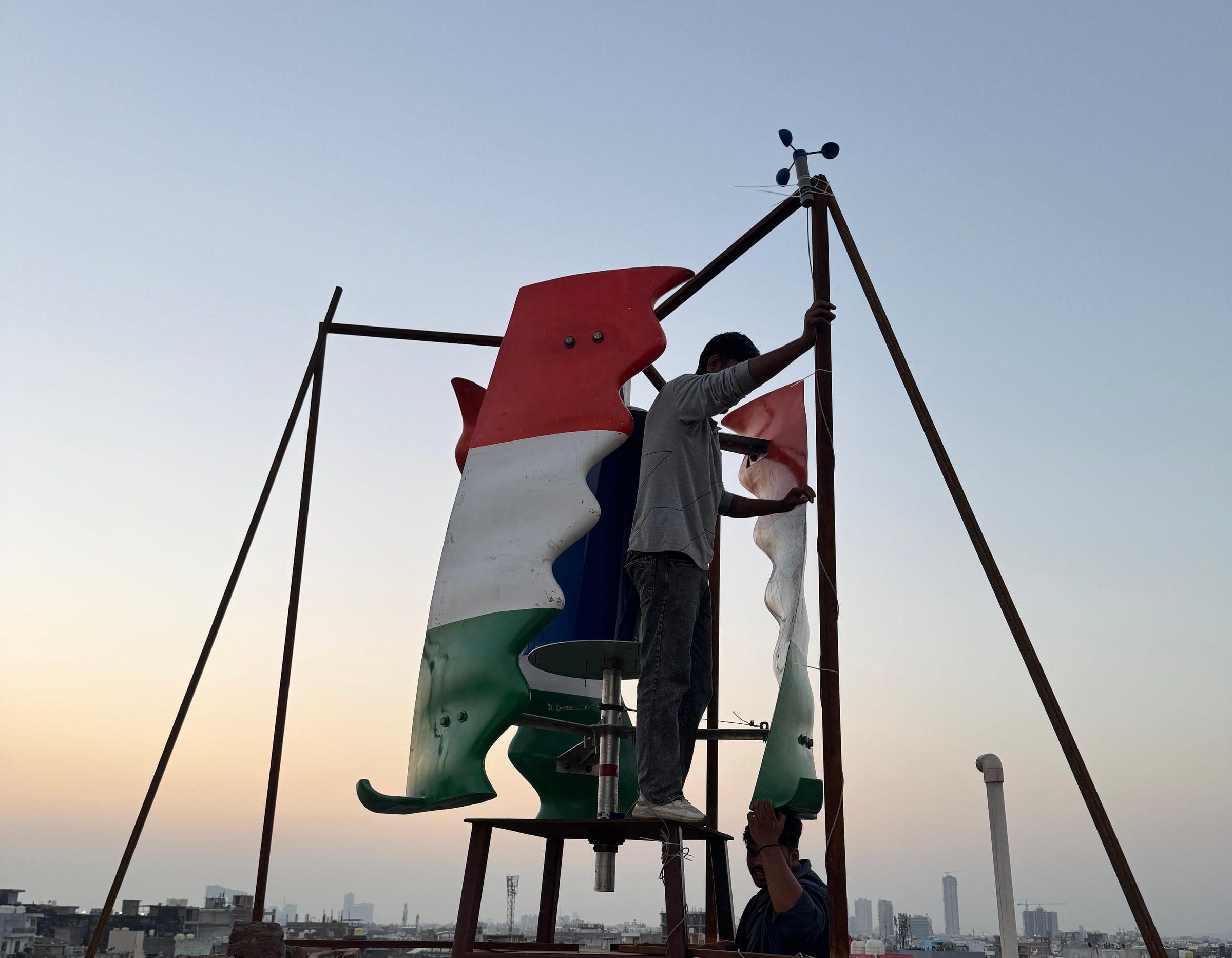 At golden hour, a crew member stands on the steel stand beside the tricolour machine, fixing a cup anemometer to the top of the mast against a pale sky.