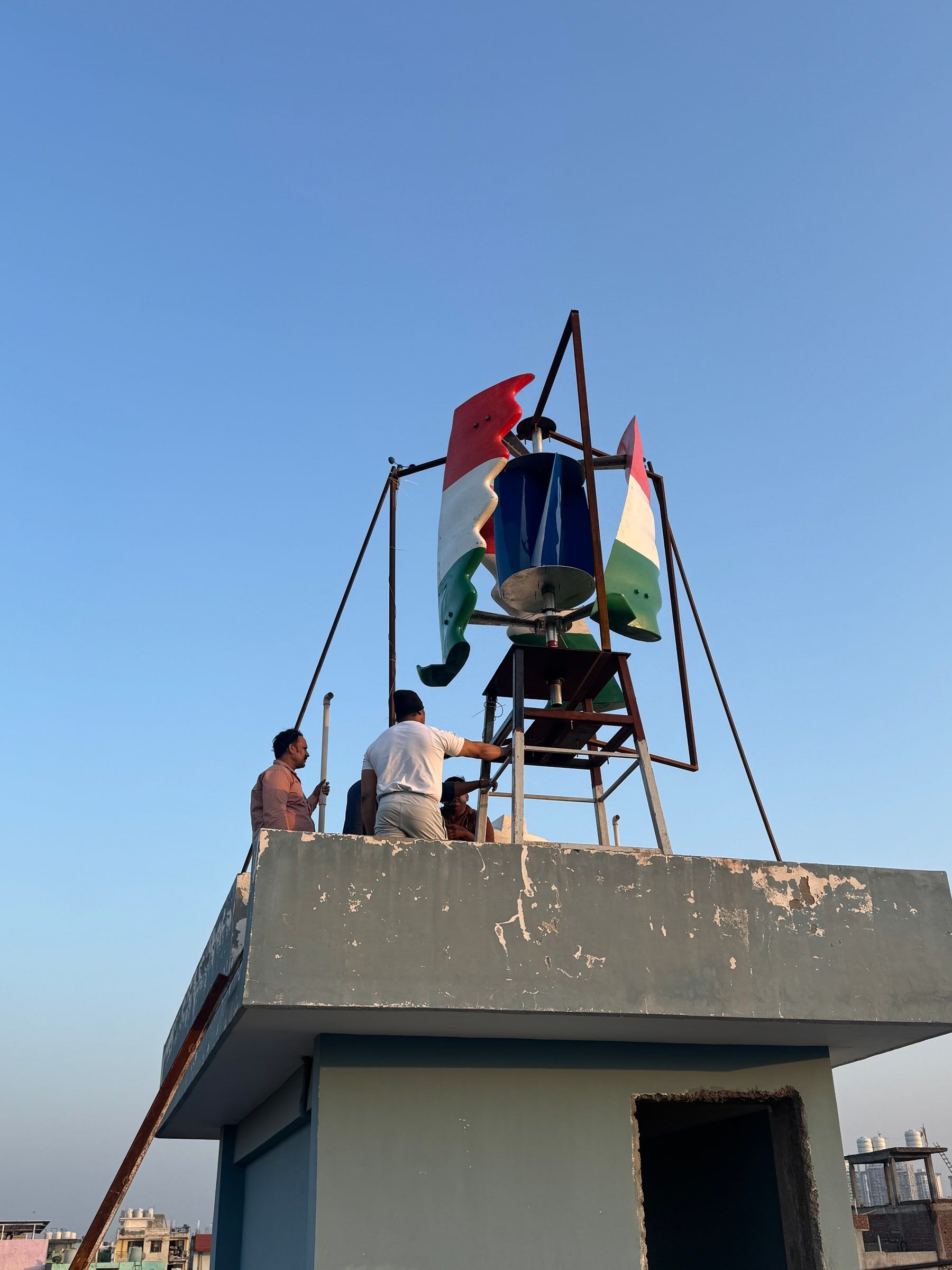 The full tricolour machine on its steel stand on a rooftop under a clear blue sky, three of the crew working at the parapet below it.