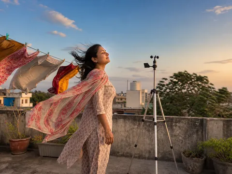 A homeowner on a breezy Indian evening terrace, washing flapping on the line, standing beside a small wind-measurement instrument.